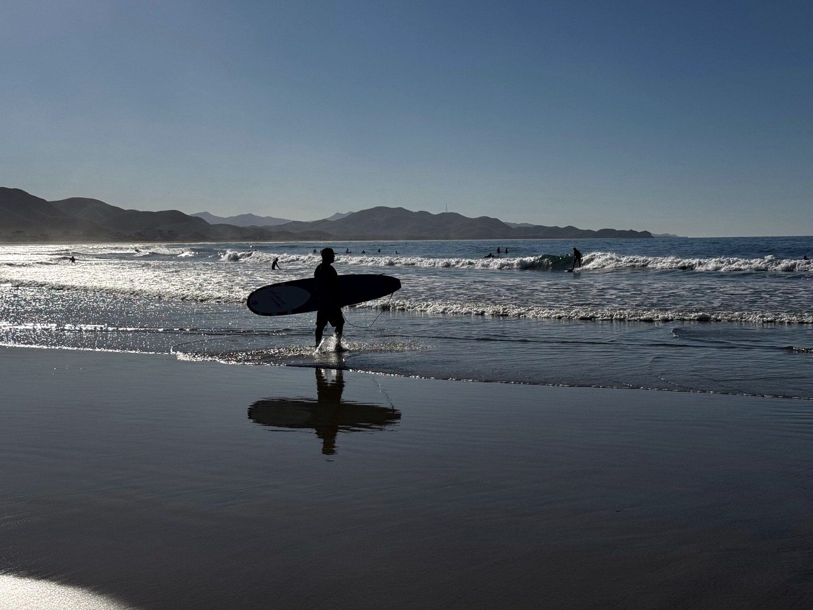 A lone surfer walking out across the wet sand at dawn, reflected in the shallows